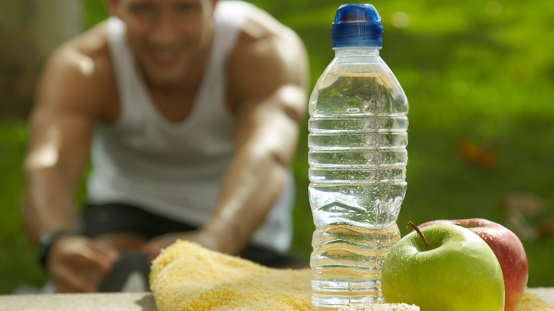 Flaša vode i jabuke u prvom planu dok muškarac u sportskoj opremi radi istezanje u prirodi