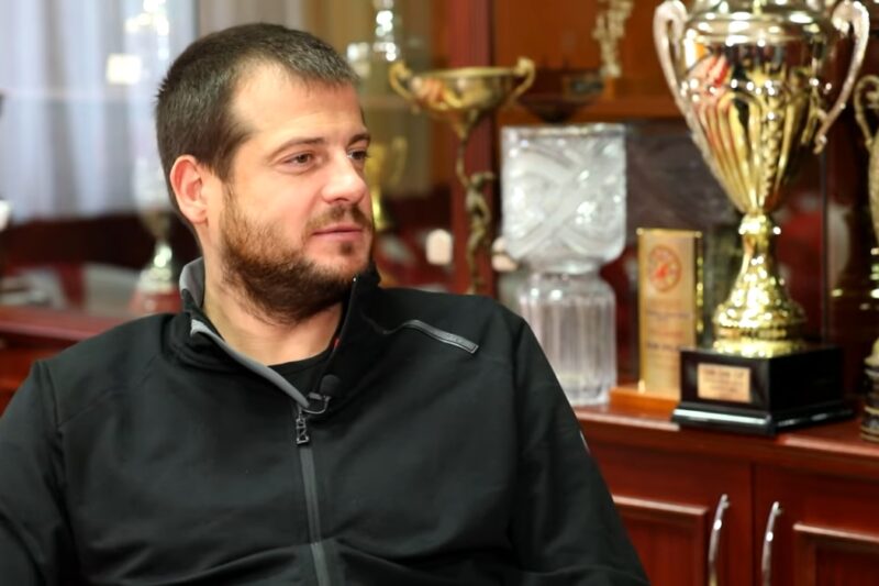 Fudbalski trener sedi u prostoriji sa trofejima u pozadini tokom intervjua